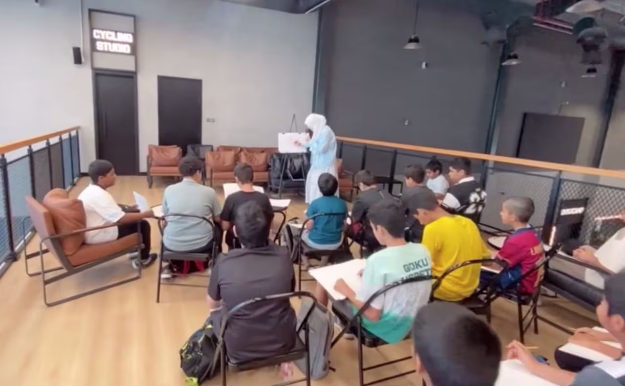 Children seated and writing while a Rawan in a blue dress stands in front of a whiteboard in a studio.