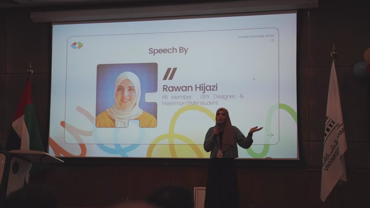 Rawan stands on stage holding a microphone and speaking, with a large screen behind her displaying a slide that reads 'Speech By Rawan Hijazi, PR Member, GFX Designer, & Freshman ITMM student,' alongside her photo.